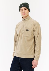Beige fleece sweatshirt met een halve rits kraag en een borstzak. Draagt een donkergroene beanie met een logo. Zachte textuur.