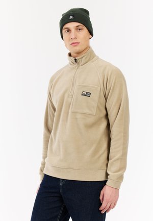 Beige fleece sweatshirt met een halve rits kraag en een borstzak. Draagt een donkergroene beanie met een logo. Zachte textuur.