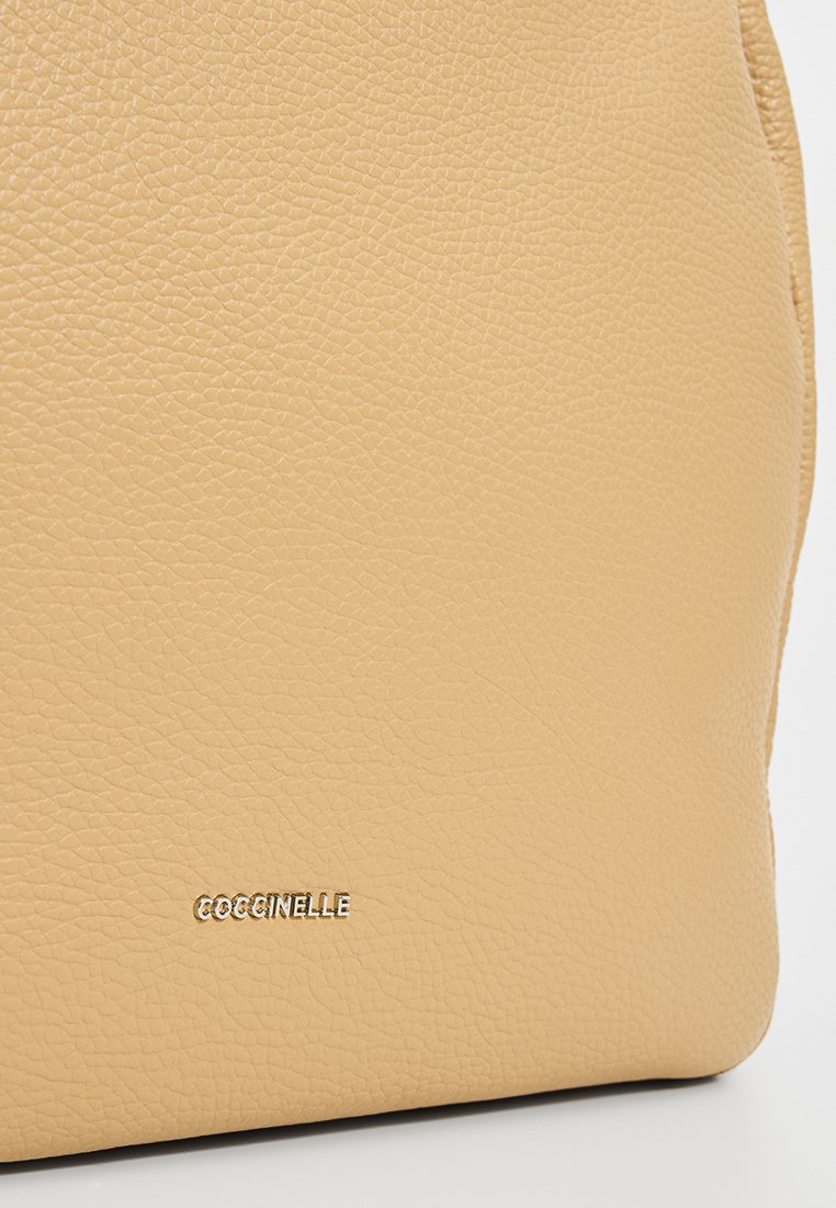 Beige teksturēta ādas rokassoma ar mazu zelta zīmola logo "COCCINELLE", iespiestu tuvu apakšējai malai.