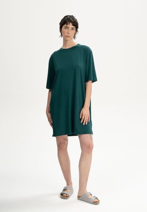 Vrouw met kort donker haar, gekleed in een losse donkergroene jurk met korte mouwen en grijze sandalen met open tenen, staand tegen een effen lichte achtergrond.