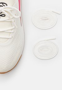 Bílé sportovní boty s texturovaným svrškem, s růžovými akcenty a dvěma páry bílých tkaniček úhledně svinutých vedle nich.