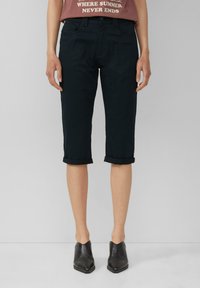 Schwarze Capri-Hosen mit umgeschlagenem Bund und glatter Textur, kombiniert mit einem taillierten braunen T-Shirt mit Grafik und schwarzen Knöchelstiefeln.