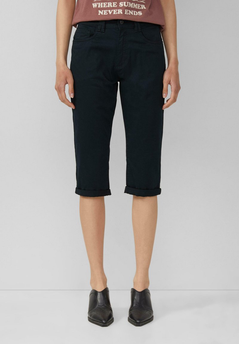 Schwarze Capri-Hosen mit umgeschlagenem Bund und glatter Textur, kombiniert mit einem taillierten braunen T-Shirt mit Grafik und schwarzen Knöchelstiefeln.