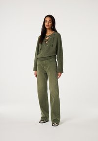 Vrouw staand, gekleed in een olijfgroene trui met veters, bijpassende wijde broekspijpen en zwart-witte schoenen met patroon, tegen een witte achtergrond.