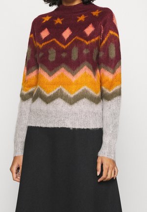 Kobieta w wielokolorowym dzianinowym swetrze z wzorami w zygzaki i geometryczne w kolorach bordowym, pomarańczowym, różowym i szarym, w połączeniu z czarną spódnicą.