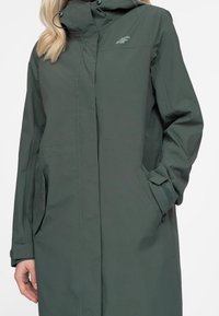 Lang grøn vandtæt frakke med hætte, fuld lynlås foran, to sidelommer og justerbare manchetter. Har et diskret logo på brystet.
