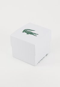 Balta kartoninė dėžutė su tekstūruota danga, turinti žalią krokodilo logotipą ant viršaus ir užrašą „Lacoste“ ant šono.