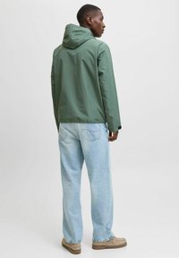 Vannavstøtende grønn jakke med hette, kombinert med lys blå jeans med avslappet passform og beige fottøy. Glatt tekstur, minimalistisk design.