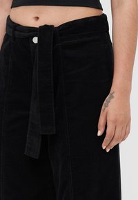 Musthave muströöpidega mustad puuvillased püksid, millel on laiad säärte lõiked. Kanga tekstuur on selgelt nähtavate soontega ja nupuga kinnitamine.