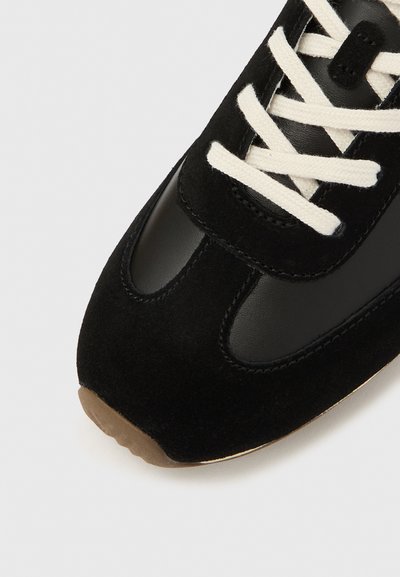 Fekete vászoncipő, amely nubukbőr és bőr anyagok kombinációját tartalmazza. Bézs színű fűzőkkel, lekerekített orral és texturált gumitalppal rendelkezik.