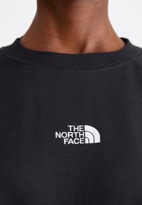 Gros plan d'une personne portant une chemise noire avec le logo blanc « The North Face » brodé sur la poitrine.