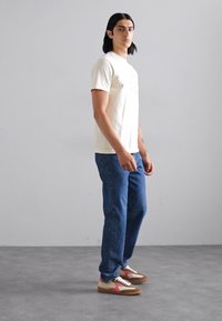 Beige short-sleeved t-shirt, blue denim jeans, and multi-color sneakers with pink accents. Model stands against a light gray background.