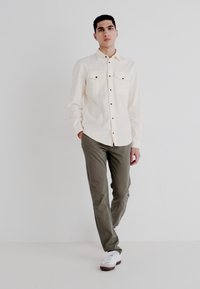 Light cream button-up shirt with two chest pockets, paired with olive green pants and white sneakers. Smooth fabric, relaxed fit, minimal design.