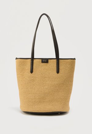 Beige kudottu olkalaukku, jossa on pitkät mustat nahkahihnat ja musta reunus, edessä pieni suorakaiteenmuotoinen logopläkki.