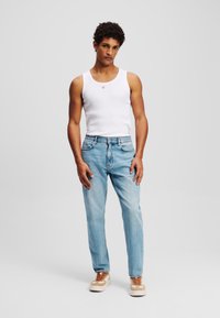 Balta berankovė marškinėliai, šviesiai mėlyni tiesaus kirpimo džinsai ir bežiniai sportbačiai su rudomis raištelėmis, stovintis neutralioje fone.