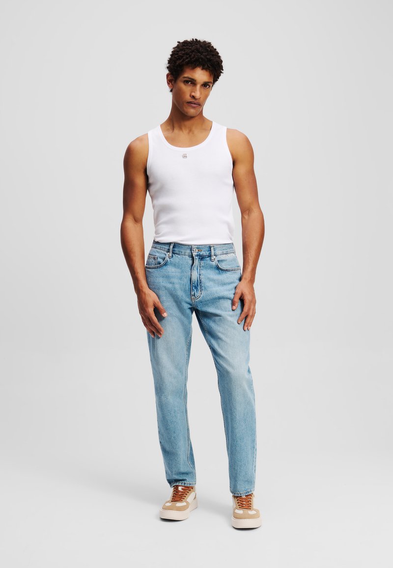 Balta berankovė marškinėliai, šviesiai mėlyni tiesaus kirpimo džinsai ir bežiniai sportbačiai su rudomis raištelėmis, stovintis neutralioje fone.