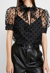Czarna bluzka w kropki z przezroczystymi bufkami, wyraźnym dekoltem w serek oraz związanym kokardą kołnierzykiem, zestawiona z spodniami z sztucznej skóry o wysokim stanie.