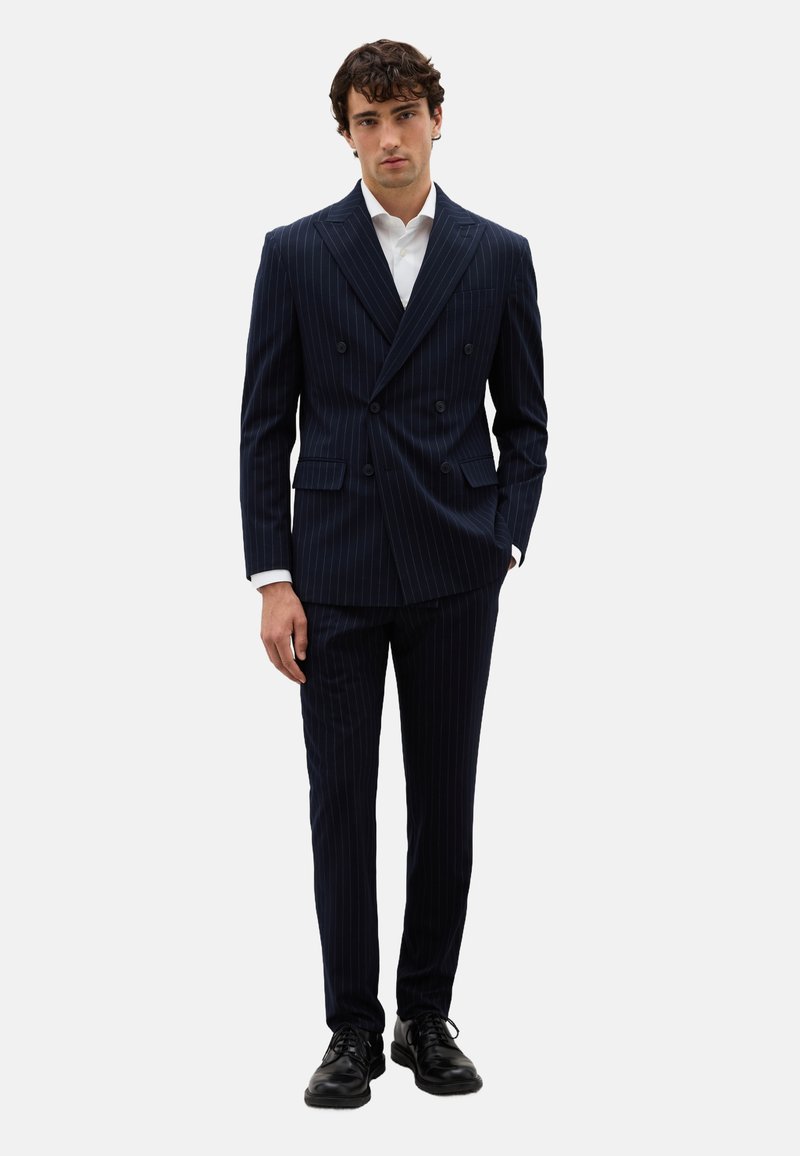 Traje azul marino de rayas diplomáticas, de corte cruzado y con solapas muesca, combinado con una camisa blanca y zapatos de cuero negros. Ajuste entallado y diseño estructurado.