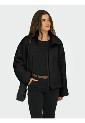 Schwarze Fleecejacke mit hohem Kragen, Frontreißverschluss, Seitentasche und weiten Ärmeln, getragen über einem schwarzen Oberteil und Hose.