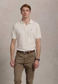Valge polo särk, millel on krae ja väike logo, koos khaki pükstega. Vöödega taljes, tekstuuriga kangas ja sobiv disain.