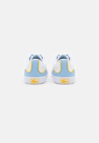 White sneakers with blue accents and yellow detailing, featuring a smooth texture and a prominent logo at the heel. Low-top design.