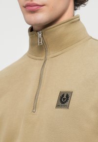 Μπεζ φούτερ με φερμουάρ, διαθέτει ψηλό γιακά, ασημένιο φερμουάρ και μαύρο κεντητό λογότυπο στο αριστερό στήθος. Ομαλή υφή βαμβακιού.