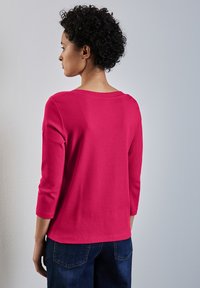 Fuchsia Ribbed-Top mit dreiviertel Ärmeln, verfügt über einen weiten Ausschnitt und eine subtile Textur. Kombiniert mit blauen Jeans.