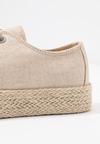 Chaussure en toile beige avec une semelle en jute tressé, forme arrondie et détails cousus le long des bords. Dessus texturé avec des œillets pour les lacets.