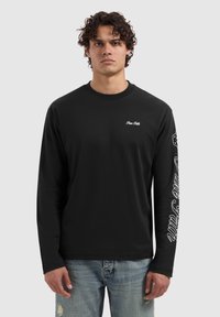Zwarte lange mouw shirt van katoen, met een klein wit logo op de borst en witte tekst langs de linkermouw. Casual pasvorm.