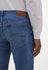 Modré džínové kalhoty z denimu s hnědým koženým štítkem na v pase s logem "Lee", detaily na zadních kapsách a světlými švy.