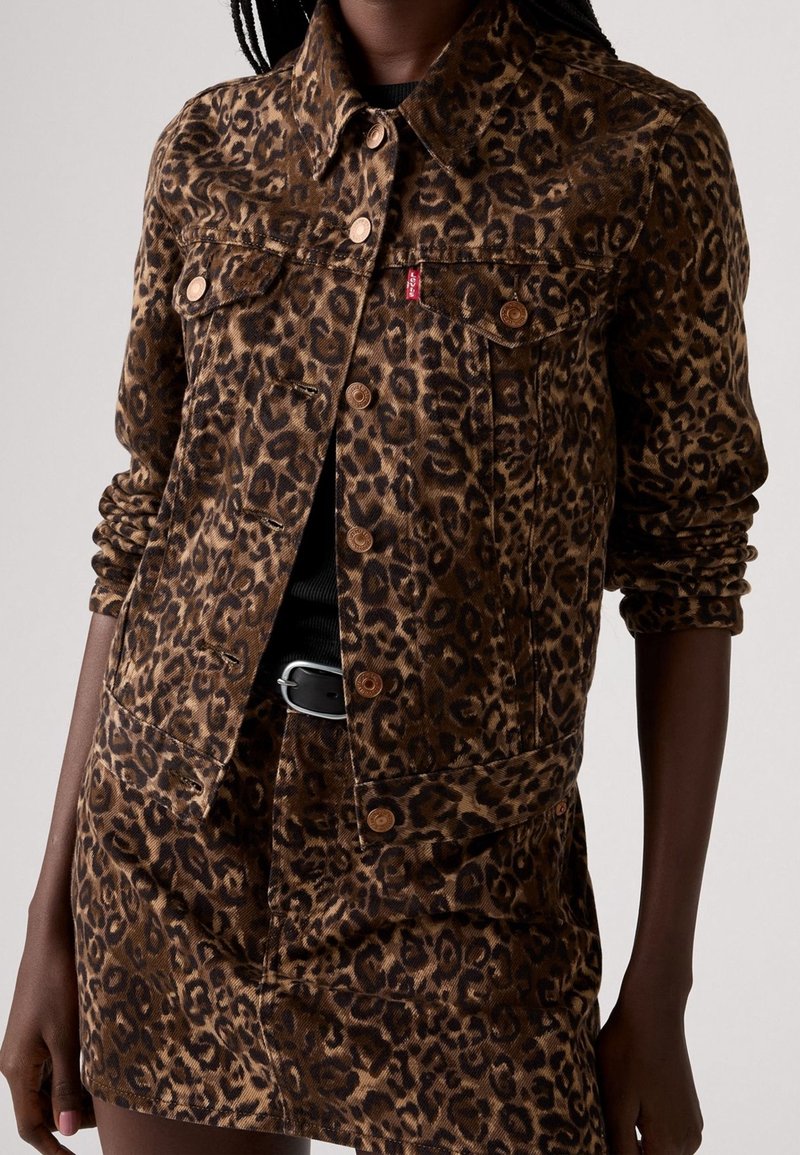 Femme portant une veste marron et noire à imprimé léopard et une jupe assortie avec des manches retroussées et une ceinture noire, sur un fond uni.