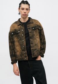 Zagorela rjava denim jakna s bledim črnim vzorcem, zapiranje na gumb, žepoma na prsih in posrnjenimi rokavi, nosi se preko črne majice.