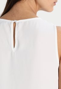 Bílá bezramínková blůza s malým trojúhelníkovým výřezem na zadní straně, s knoflíkovým zapínáním nad výřezem. Hladký povrch a minimalistický design.