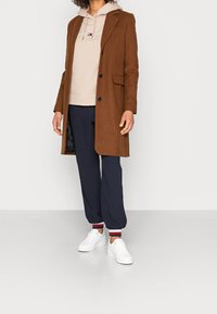 Persona con un abrigo marrón abotonado sobre una sudadera beis, pantalones de chándal azul marino con puños a rayas y zapatillas blancas, de pie sobre un suelo gris.