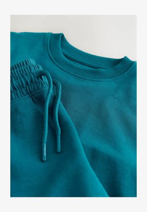 Tirkīzzils pelēks sporta krekls un atbilstošas bikses, ar ribotām aprocēm un jostasvietu, aukliņām un gludu auduma tekstūru.