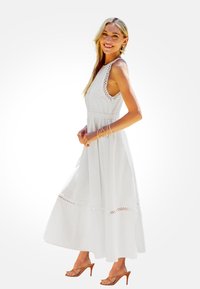Biała maxi sukienka o bezrękawowym kroju, z koronkowymi akcentami na ramionach i u dołu, z wcięciem w talii oraz miękkim, lekkim materiałem. Zestawiona z brązowymi szpilkami.