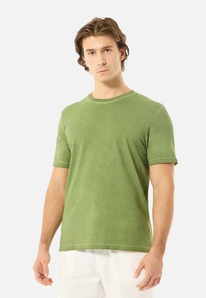 Jeune homme aux cheveux châtain clair portant un t-shirt vert uni à manches courtes et un pantalon blanc, debout devant un fond clair.