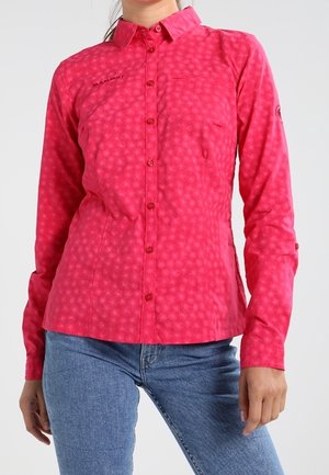 Frau trägt ein langärmliges, rosa gemustertes Hemd mit Knöpfen und blaue Jeans, steht mit einer Hand seitlich am Körper.