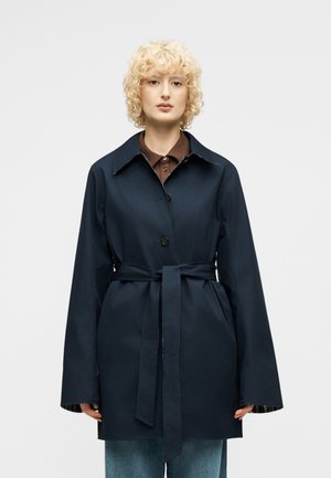 Blondynka o kręconych włosach, ubrana w granatowy płaszcz z paskiem, na brązowej koszuli i niebieskich dżinsach, zwrócona przodem na jednolitym tle.