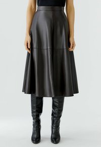 Vrouw in een donkerbruine leren midi-rok met hoge taille en zwarte, leren laarzen tot de knie, staand tegen een effen achtergrond.