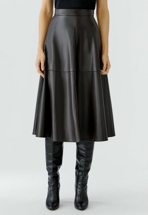 Vrouw in een donkerbruine leren midi-rok met hoge taille en zwarte, leren laarzen tot de knie, staand tegen een effen achtergrond.