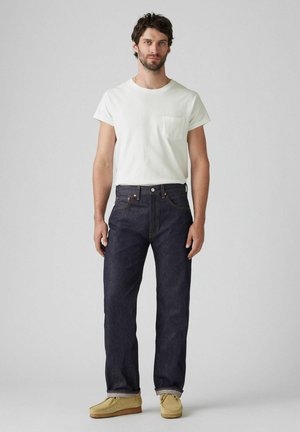 Jeans in denim blu scuro con taglio dritto, abbinati a una camicia bianca a maniche corte con taschino, e scarpe slip-on beige.