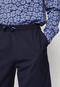 Personne portant un pantalon taille élastique bleu marine avec cordon et une chemise bleue à fleurs à manches longues, main dans la poche.