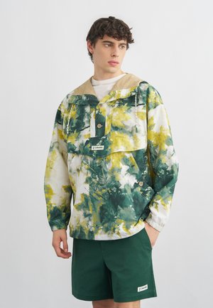 Mladi moški v zeleni, rumeni in beli pulover jakni s kapuco in temnozelenih kratkih hlačah, stoji pred enobarvnim svetlim ozadjem.