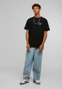 Sort bomulds T-shirt med hvid tekst "Dead WRONG" på brystet, parret med lyseblå brede jeans og sort-hvide sneakers.