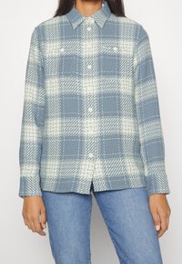 Jasnoniebieska koszula w kratę, uszyta z teksturowanej tkaniny. Posiada zapięcie na guziki, kołnierz oraz dwie kieszenie na piersiach z białym wzorem przeszyć.