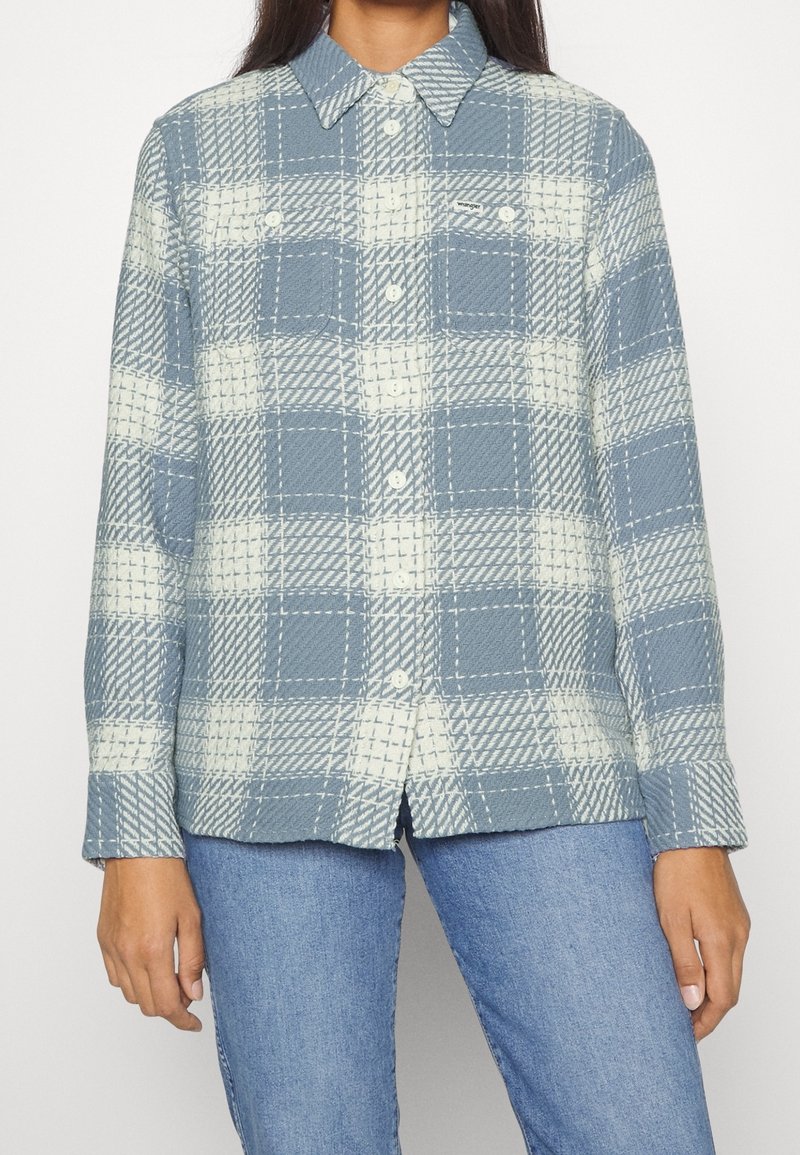 Jasnoniebieska koszula w kratę, uszyta z teksturowanej tkaniny. Posiada zapięcie na guziki, kołnierz oraz dwie kieszenie na piersiach z białym wzorem przeszyć.