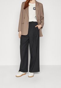 Braun blazer mit doppelter Knopfleiste, weißes T-Shirt mit Logo, schwarze weit geschnittene Hose und weiße Sneaker mit braunen Sohlen.