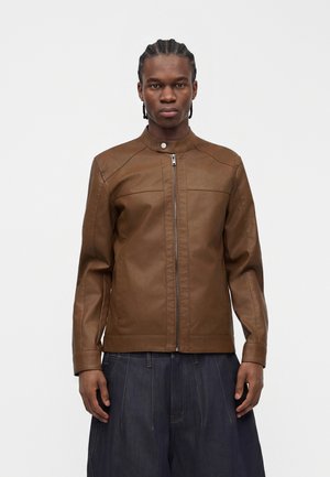 Homme aux cheveux tressés portant une veste en cuir marron zippée et un pantalon en jean sombre ample, debout devant un fond uni.