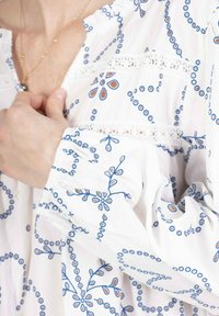 Blouse blanche avec des motifs floraux bleus et oranges, dotée d'accents en dentelle et de manches bouffantes. Tissu doux avec une coupe ample.